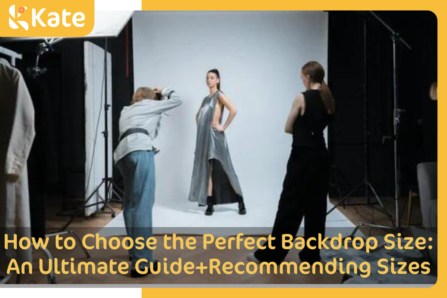 A photographer is taking pictures of a female model in front of a white backdrop, another model stands at side of the photogrpher.