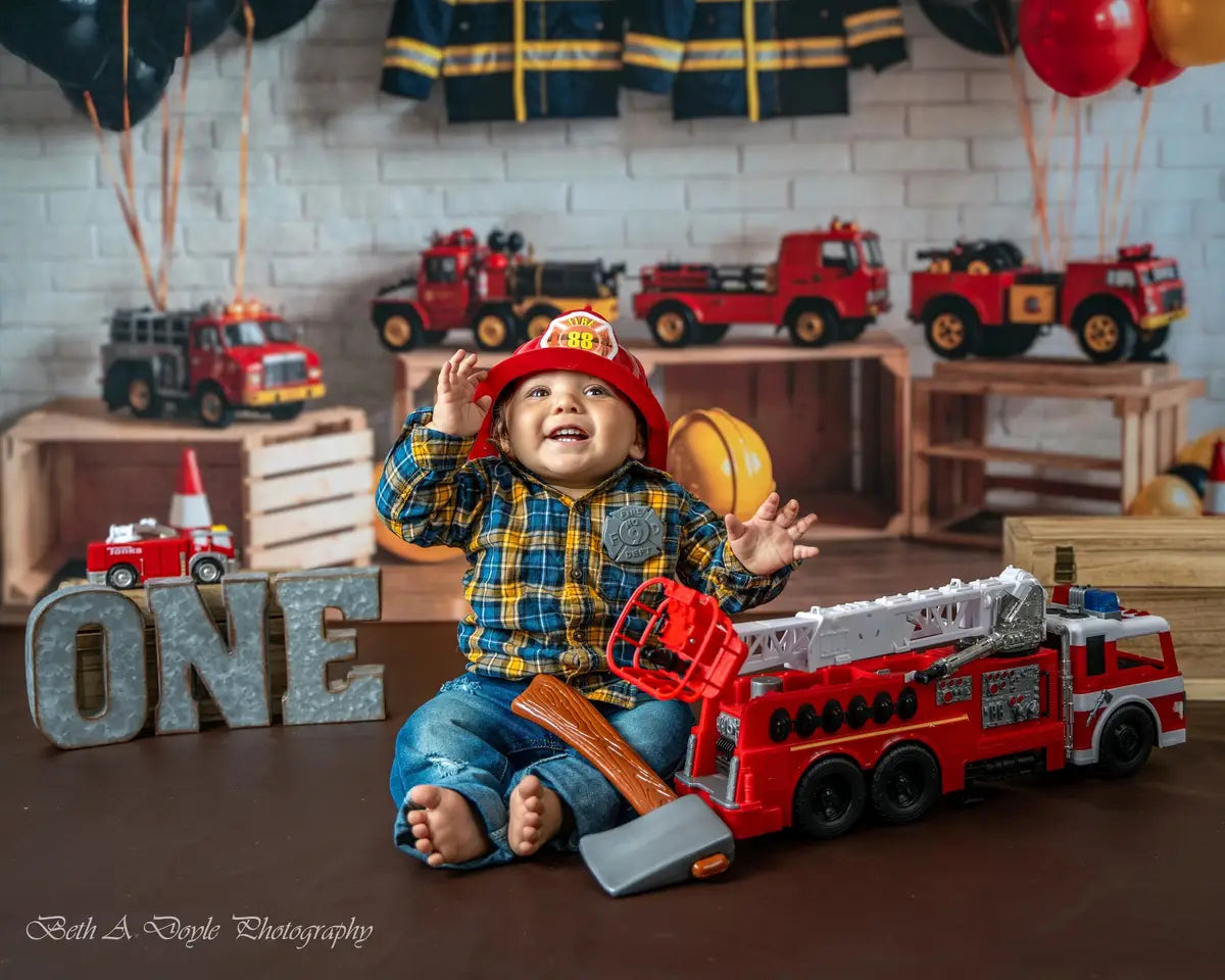 Tema de lucha contra incendios en cumpleaños con globos coloridos, juguete de auto y uniforme, todo diseñado por GQ en un fondo de pared de ladrillo blanco.