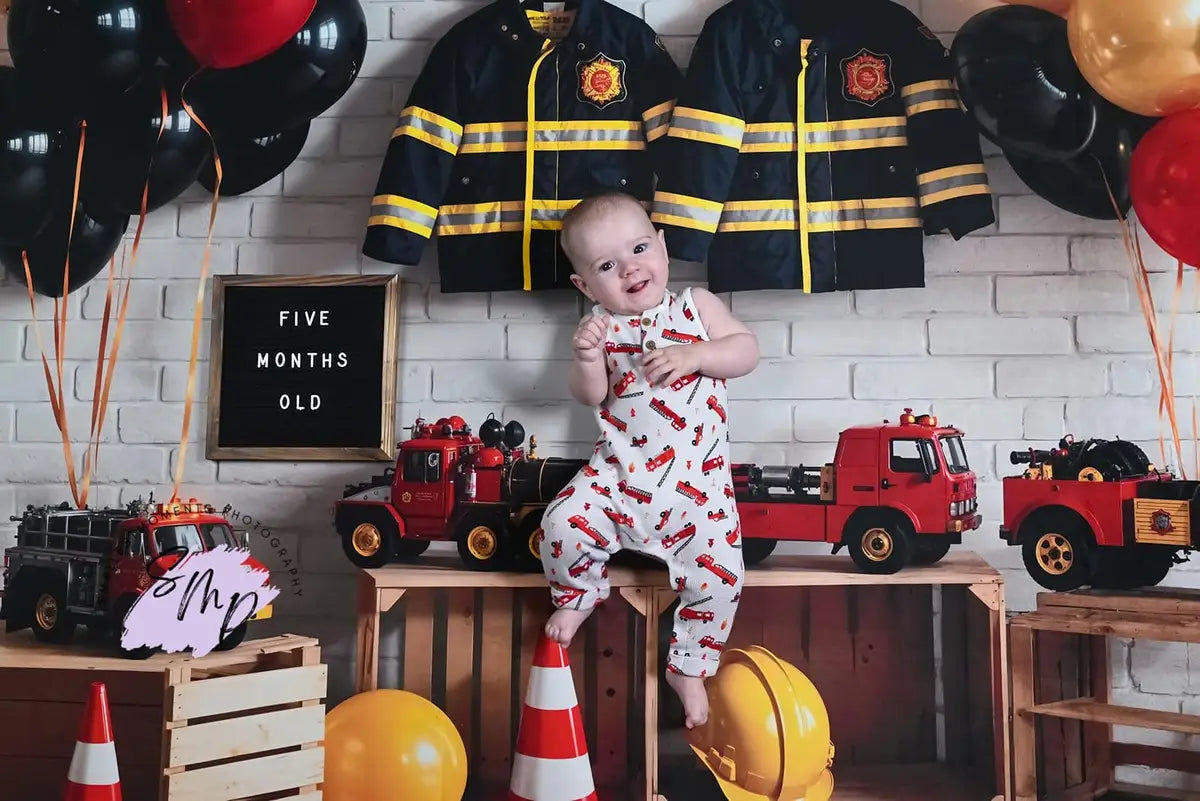 Tema de lucha contra incendios en cumpleaños con globos coloridos, juguete de auto y uniforme, todo diseñado por GQ en un fondo de pared de ladrillo blanco.